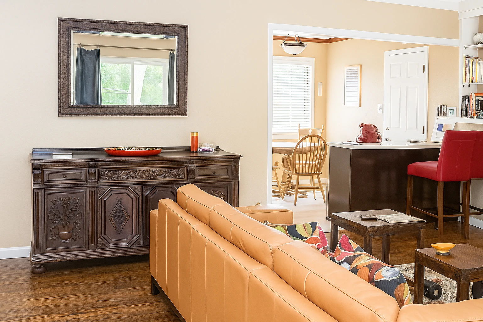 Recreate this interior photo with realistic daylight exposure and tonal balance suitable for professional real estate photography. Simulate natural sunlight coming from the window at the left, filling the room evenly with soft ambient light. Adjust exposure so that the wall tones remain light beige with visible surface texture, and the white trim stays neutral white without glare or flash. Reduce warm color cast from the walls by slightly cooling the white balance, while maintaining a gentle warmth in wooden furniture. Balance highlights and shadows with smooth tonal transitions—no harsh contrast or clipped whites. Ensure reflective surfaces such as the wood cabinet and mirror frame show soft, diffused reflections instead of sharp glare. Preserve fine texture on the sofa fabric and floor. Apply subtle HDR-style tone mapping so both indoor and outdoor areas are visible naturally. The final image should feel bright, neutral, and professionally shot, like an MLS-ready daylight photo.